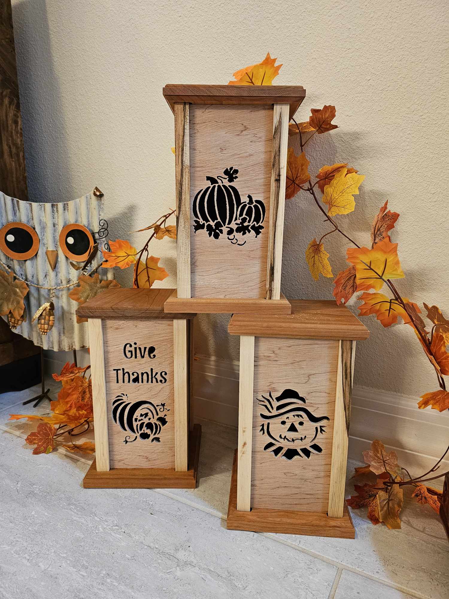 Three wooden lanterns with lantern faces depicting pumpkins, a scare crow and a cornocopia with the phrase "Give Thanks". Natural finish.