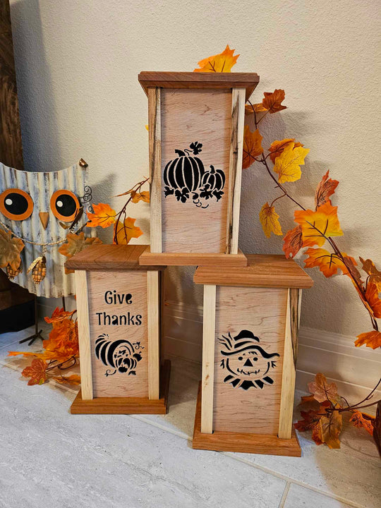 Three wooden lanterns with lantern faces depicting pumpkins, a scare crow and a cornocopia with the phrase "Give Thanks". Natural finish.