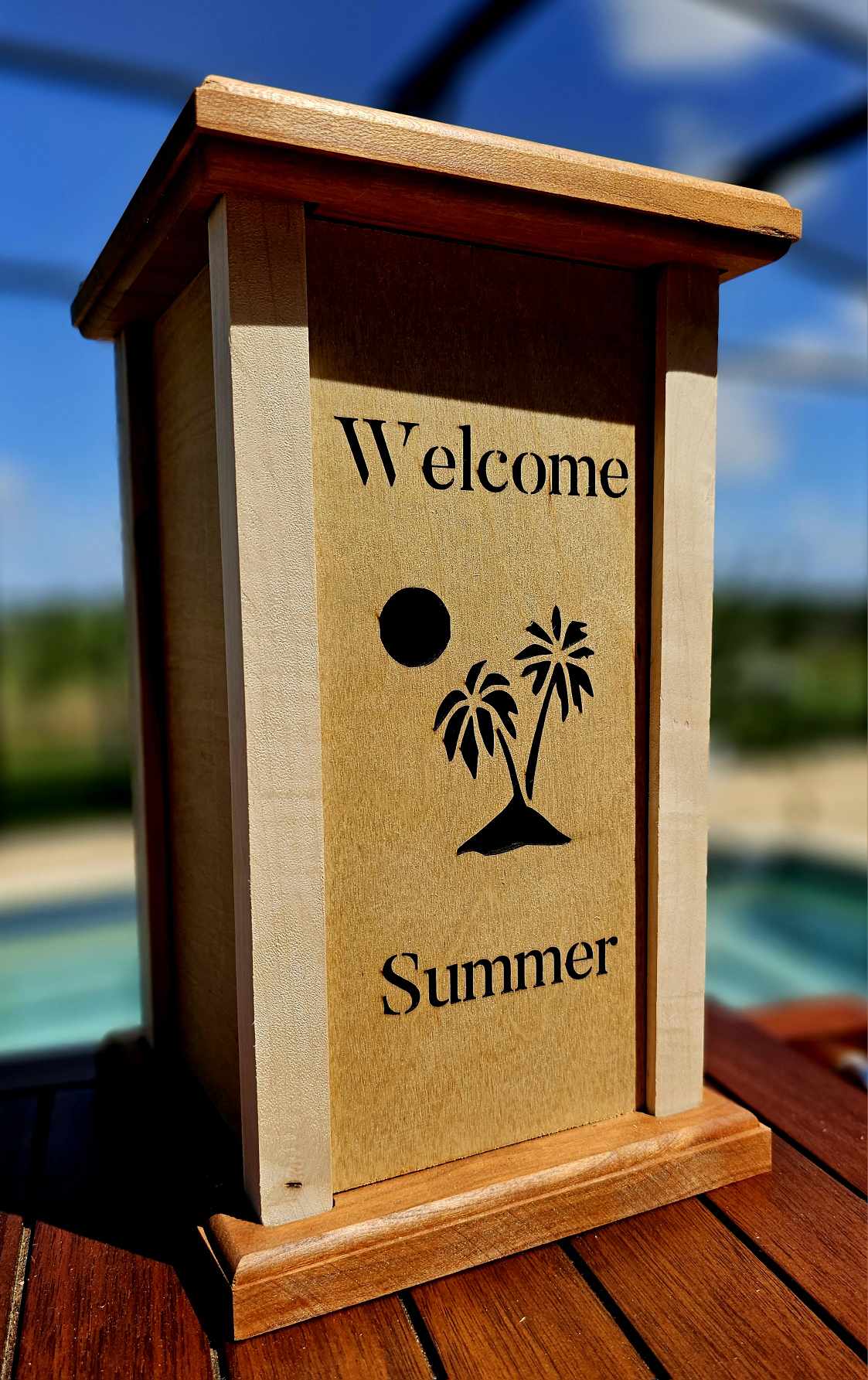 Wooden lantern face showing a island with palm trees and setting sun with the words Welcome Summer.